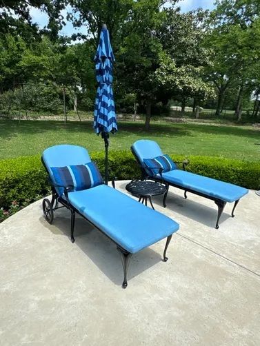 Two blue lounge chairs and a small table under a blue and white striped umbrella on a concrete patio by a grassy lawn.