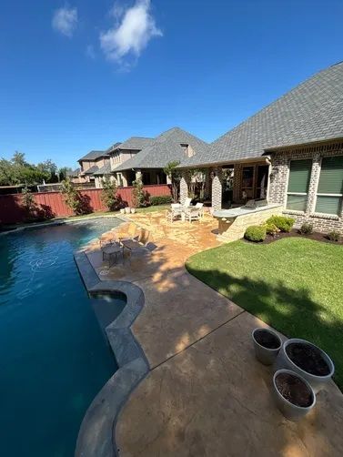 A backyard swimming pool with a tan concrete patio, outdoor seating area, and a stone-trimmed house under a blue sky.