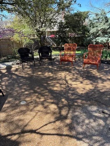 Two black chairs and two rust-colored wicker chairs sit on a gravel patio outdoors surrounded by trees.