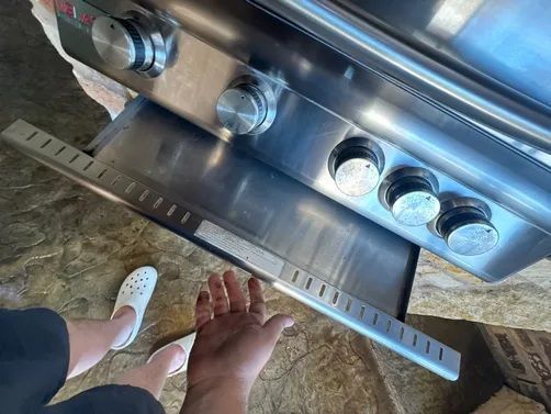 A person's hand reaches toward an open, stainless steel grease tray on a grill, with their feet visible in white crocs.
