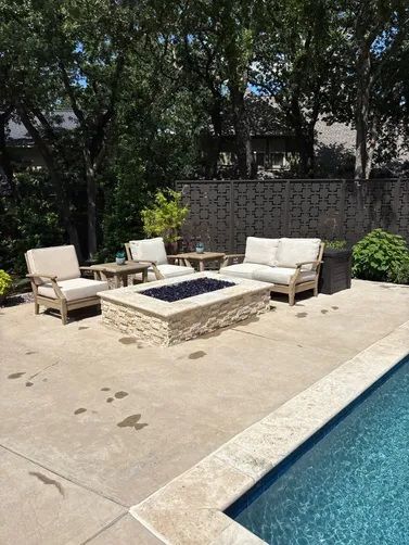 A stone fire pit surrounded by outdoor patio furniture on a concrete deck next to a backyard swimming pool.