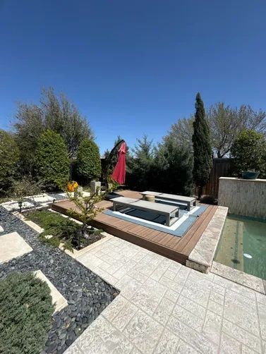 A sunny backyard patio features light-colored pavers, a wood-decked seating area with chairs, and a small pool edge.