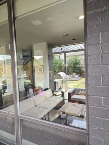 A view through a window into a covered patio with a light-colored sectional sofa, a coffee table, and an outdoor kitchen.