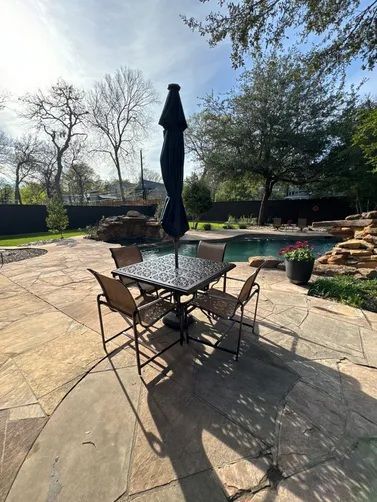 A patio set with a closed umbrella and four chairs on a stone-paved pool deck with a rock waterfall in the background.