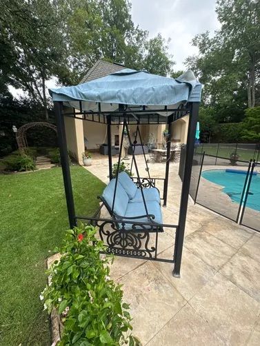 A blue outdoor porch swing with a canopy, positioned on a stone patio beside a swimming pool and a lush green lawn.