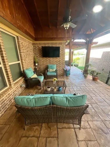An outdoor covered patio with brick walls, a mounted television, wicker furniture with turquoise cushions, and a ceiling.