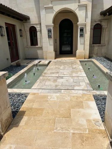 A limestone walkway flanked by two reflecting pools with small fountains leads to the arched entryway of a stone house.