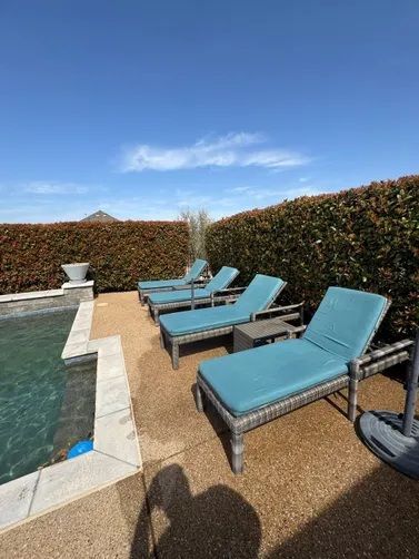Three blue lounge chairs sit on a gravel patio next to a swimming pool, bordered by a tall hedge against a blue sky.