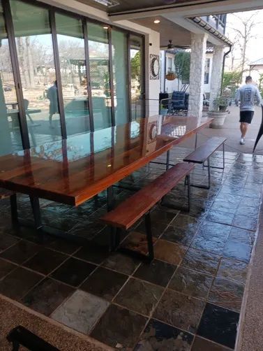 A long, polished wooden dining table with matching benches on a tiled outdoor patio, with a person walking in the distance.