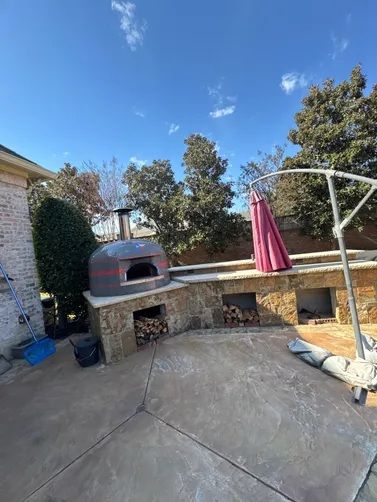 Outdoor patio with a grey pizza oven on a stone counter, featuring built-in wood storage shelves and a pink umbrella.
