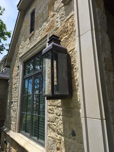 An outdoor lantern-style wall light mounted on a tan, textured stone wall next to a dark-framed window.