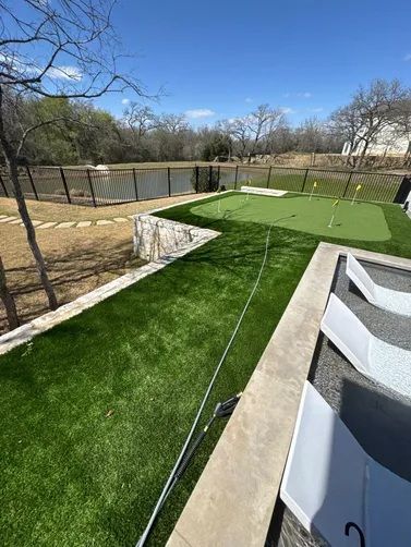 A putting green with small flags next to a pool deck with lounge chairs, overlooking a pond on a sunny day.