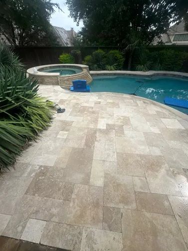 A backyard swimming pool and stone spa with a beige-toned paver deck, lush green landscaping, and trees.