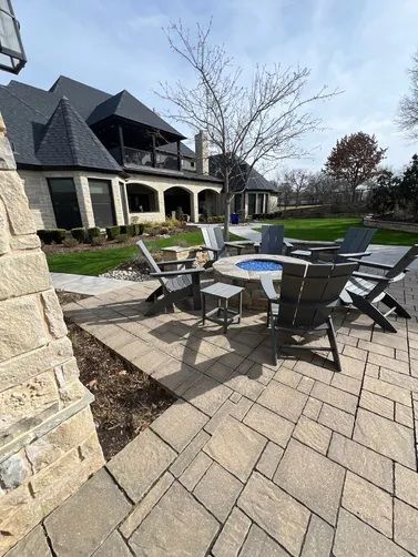 A stone patio with dark Adirondack chairs around a fire pit in front of a multi-story stone house on a sunny day.