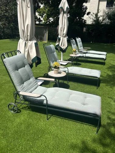 Three grey cushioned lounge chairs with small tables and closed umbrellas set on a green lawn.