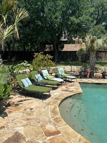 Lounge chairs on a stone patio beside a swimming pool, with lush green trees and landscaping in the background.