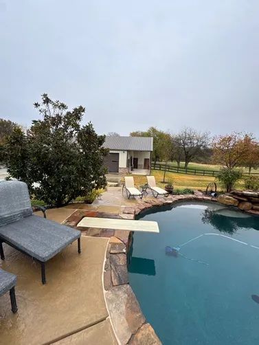A swimming pool with a diving board next to a patio with lounge chairs, overlooking a building and field on a cloudy day.
