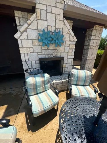 A stone fireplace on a patio with two striped chairs and a metal table under a sunny sky.