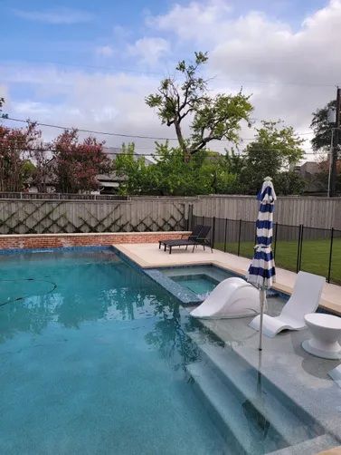 A backyard swimming pool with a sun shelf featuring white lounge chairs, an umbrella, and a wooden deck with a lounge chair.