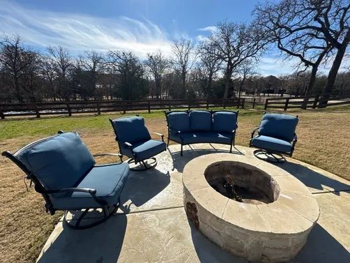 A patio with a stone fire pit surrounded by a blue sofa and three matching swivel chairs on a sunny day.