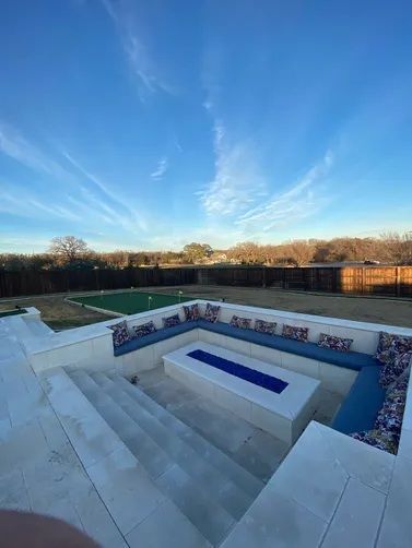 A sunken stone fire pit area with blue cushions and a rectangular gas fire feature under a bright, sunny blue sky.