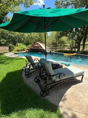 Two lounge chairs with cushions under a large green umbrella next to a swimming pool with a slide and waterfall.