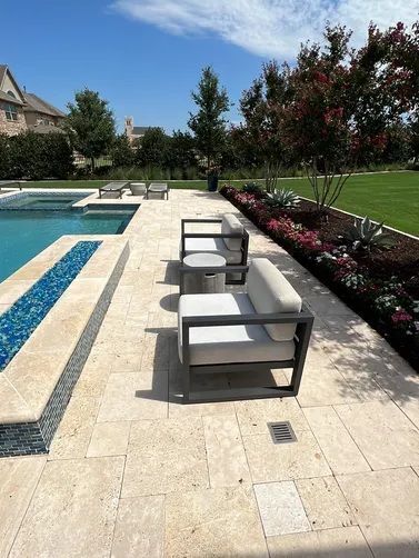 Two grey chairs with light cushions sit on a light-colored stone patio next to a swimming pool with a blue tile border.