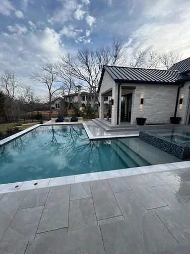 A modern stone home with a metal roof overlooks a backyard pool and spa on a bright, partly cloudy day.