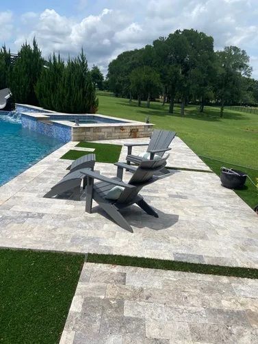 Two grey Adirondack chairs placed on a stone pool deck near a swimming pool and spa, set against a grassy landscape.