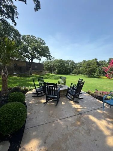 Five black rocking chairs arranged in a circle around a fire pit on a stone patio overlooking a green lawn and trees.