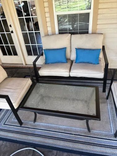 Outdoor patio set with a glass-topped table and a beige loveseat featuring two bright blue throw pillows on a porch.