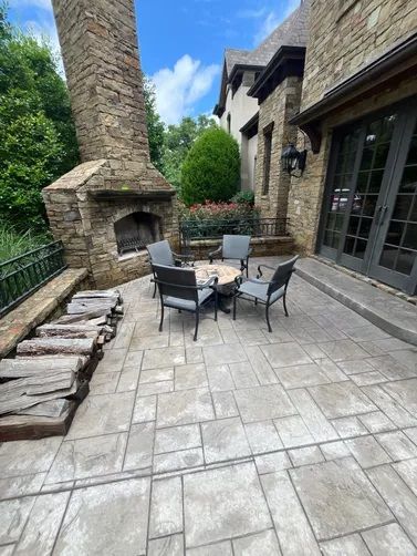 A stone patio with a fireplace, a fire pit, four chairs, and a stack of firewood against a stone house exterior.