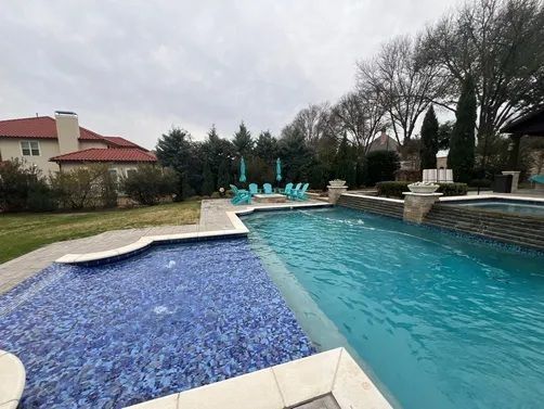 A modern swimming pool with blue tiles, a stone wall with water features, and a seating area with patio chairs on a lawn.