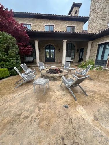A stone patio with a central fire pit surrounded by outdoor chairs in front of a multi-story stone house.