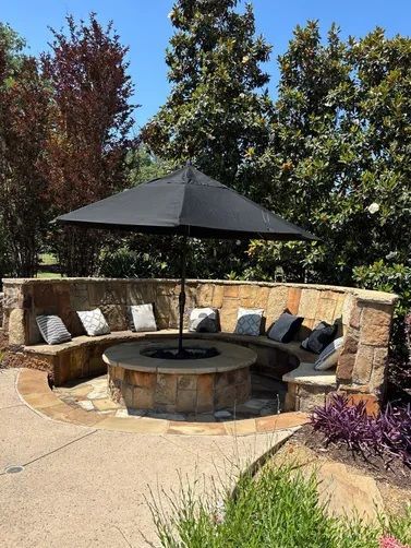 A curved stone bench with cushions circles a central fire pit shaded by a black umbrella in a sunny garden.