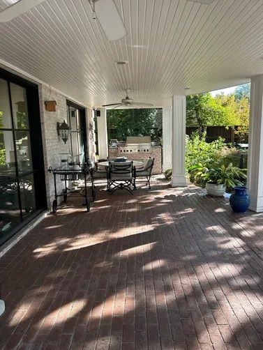 A covered brick patio with a dining table, chairs, an outdoor grill, and a ceiling fan, overlooking a lush garden.