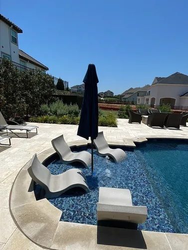 A sunny poolside scene with several light gray lounge chairs submerged on a shallow tanning ledge around a dark umbrella.