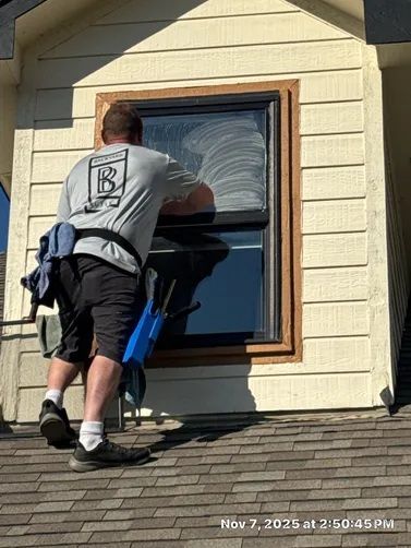 A professional cleaner stands on a shingled roof, scrubbing the top pane of a tan house window with a blue tool.