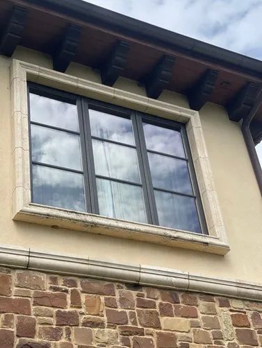 A dark-framed, multi-pane window with stone trim, set into a beige stucco and stone exterior wall under a wooden overhang.