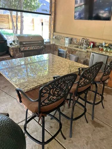 An outdoor patio featuring a granite-topped square table with three ornate metal bar stools, a built-in grill, and a TV.