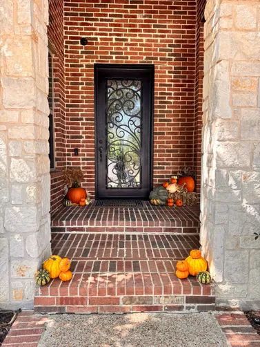 A brick front porch with stone pillars, a dark ornate glass door, and scattered autumn pumpkins on the steps.