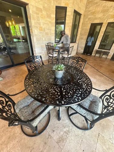 A round, ornate black metal patio table with four chairs and a small plant on a stone-tiled outdoor terrace.