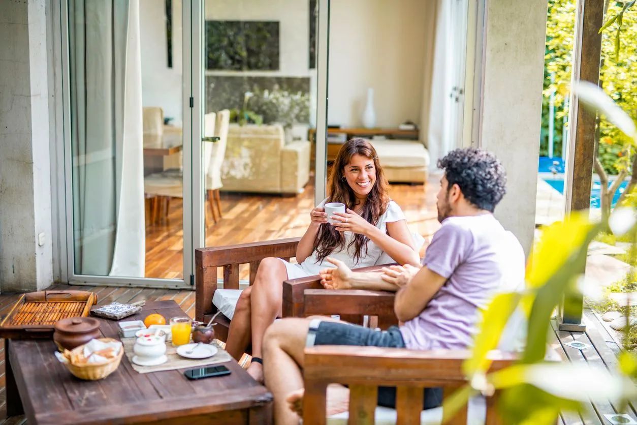 Two people talk while sitting in outdoor lounge chairs on a patio with a breakfast spread on a wooden table.