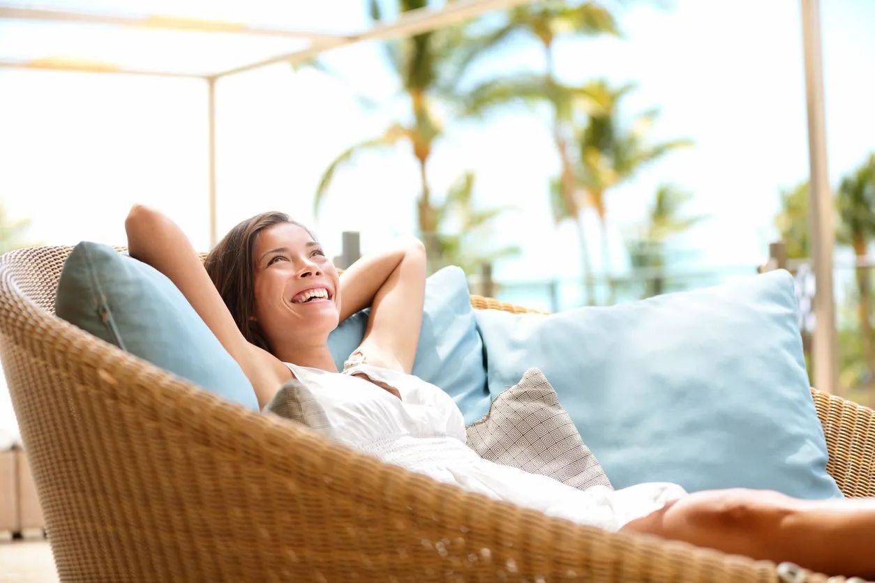 A person relaxes in a wicker chair with blue cushions, smiling while looking toward palm trees and a sunny, outdoor setting.