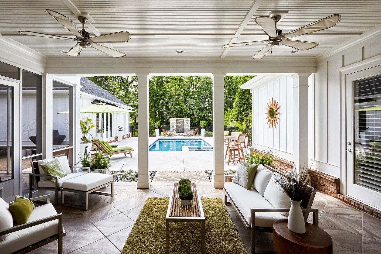 Covered patio seating area overlooking a backyard pool and greenery, with white furniture and ceiling fans.
