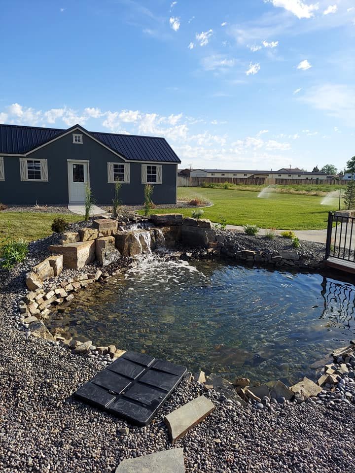 A small pond with a waterfall in front of a gray house on a sunny day.