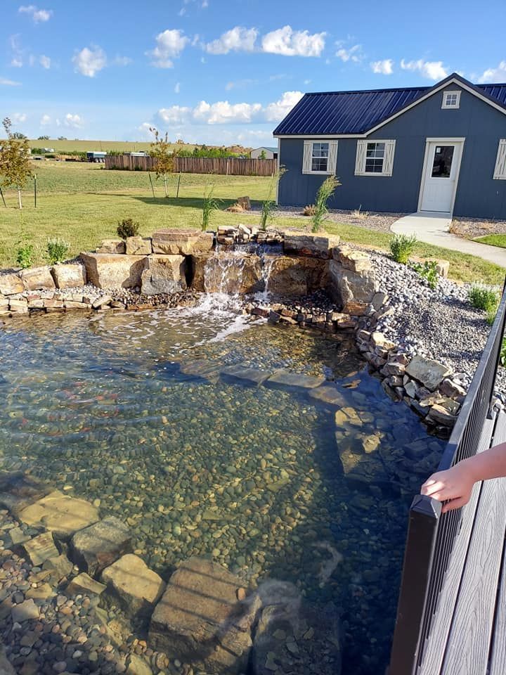 A small pond with a waterfall in front of a blue house with a black roof, sunny day.