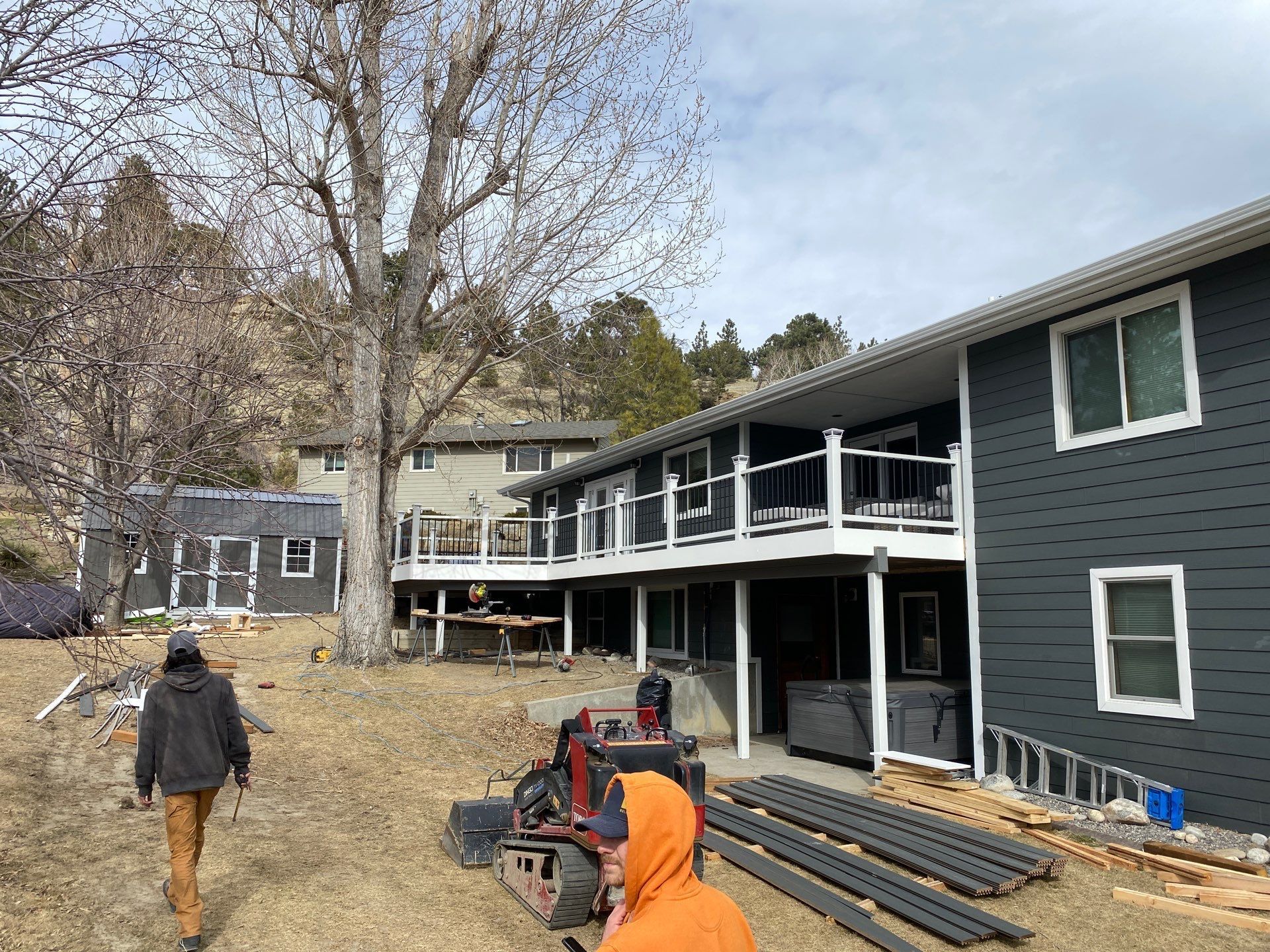 Two men near a house with gray siding and a deck; construction supplies and a small excavator.
