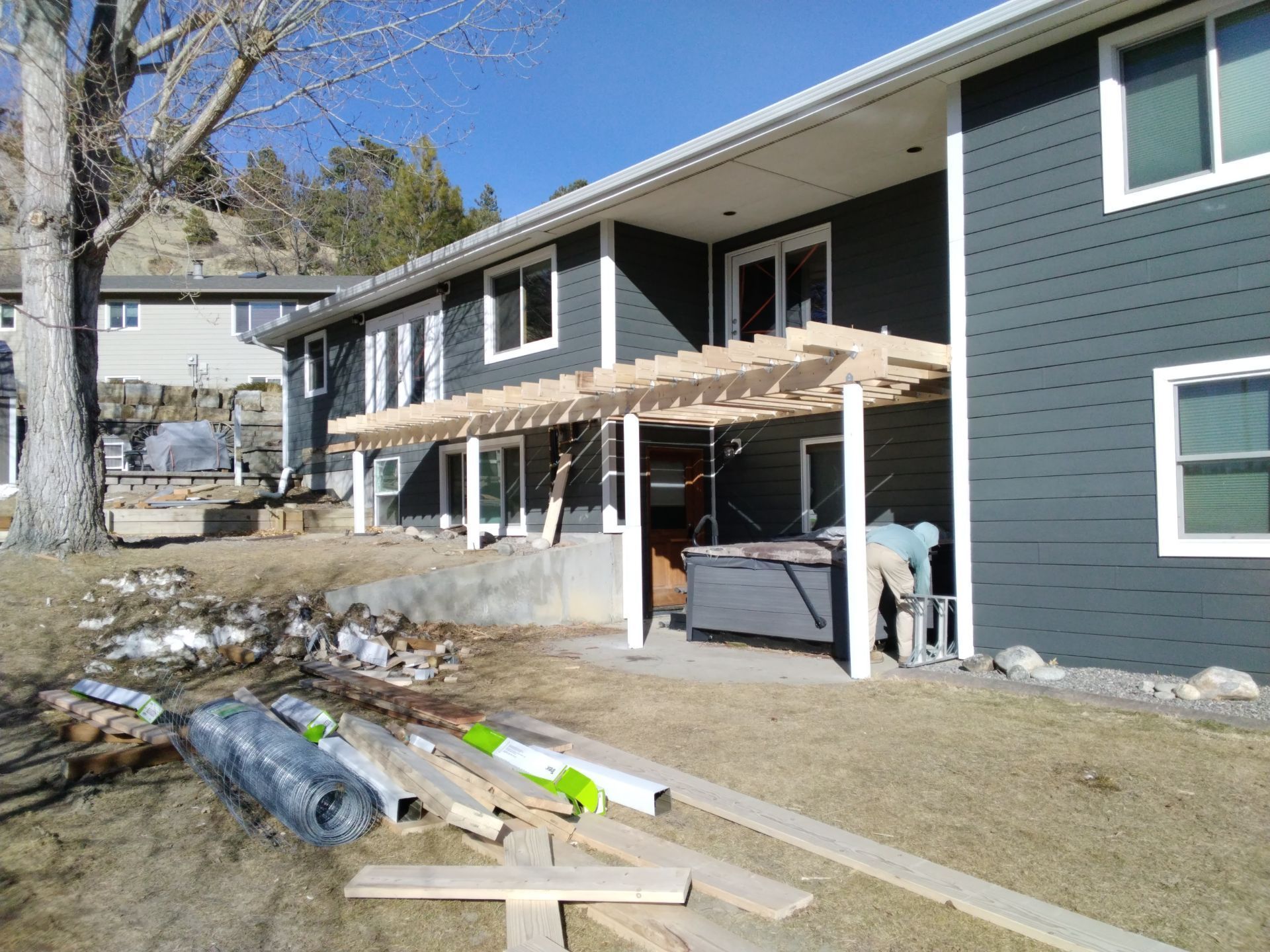 A house with a pergola over a hot tub. Construction materials lie on the ground. A person works on the structure.