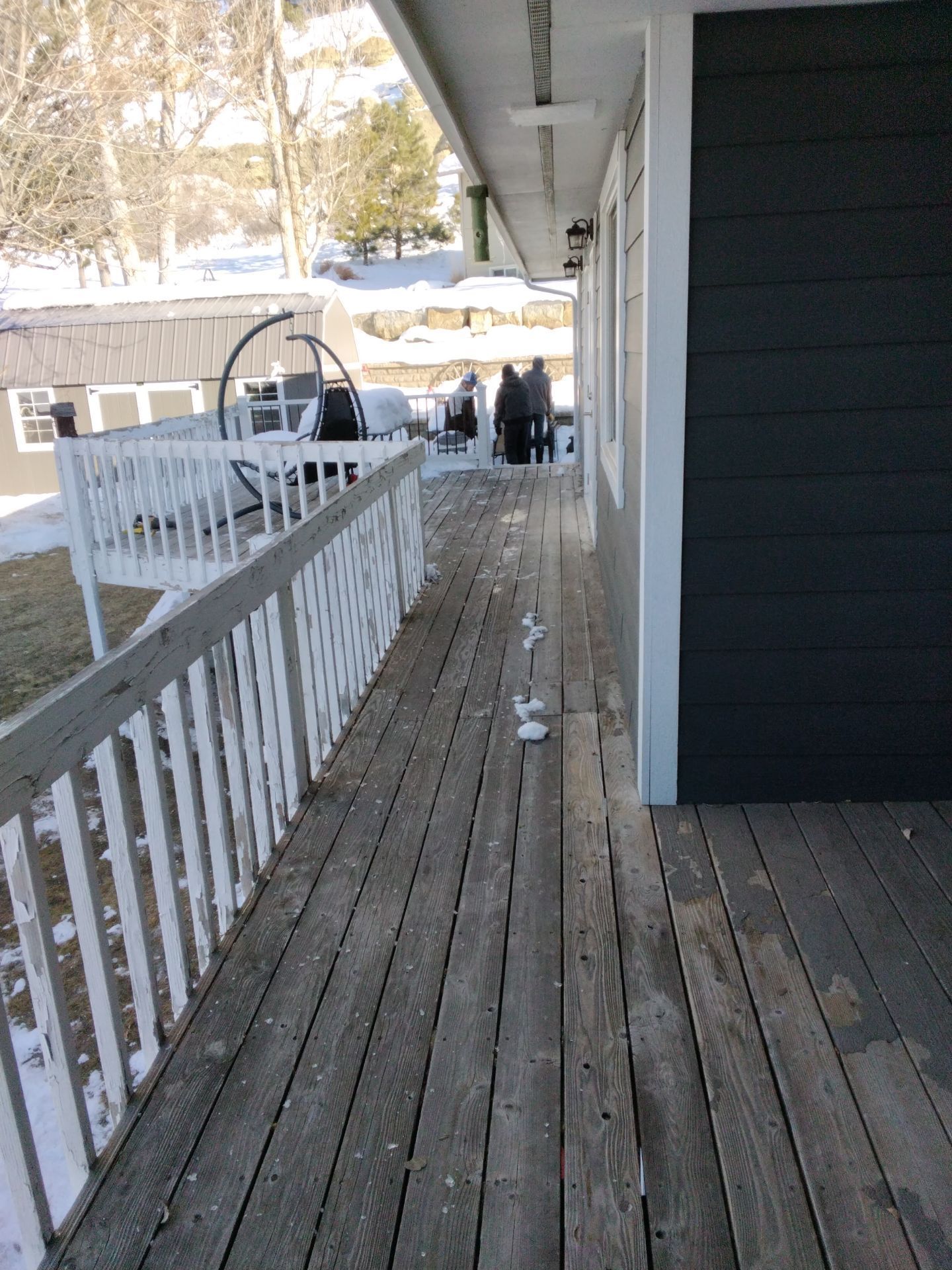 Wooden deck with white railing, leading to people standing in the distance near snow-covered ground.